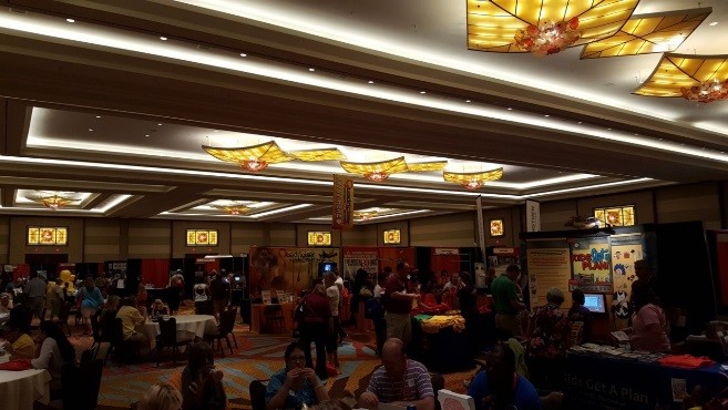 Many people stand and sit and are scattered around a large room that has many display posters standing up on tables