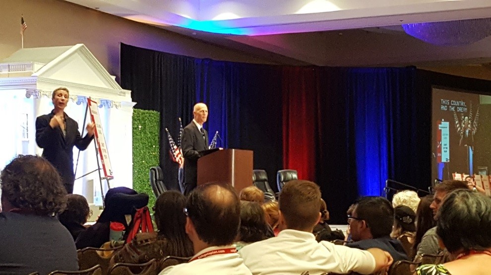 Governor Rick Scott speaks to a crowd from behind a podium. A sign language interpreter signs to his right.