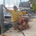 Raymond Bishop, an 84 year old man, kneeling at a dock with sailboats behind him, with his two dogs, ranger and roxie at his knees