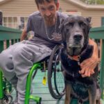 Albert Schaw, a man in a bright green manual wheelchair wearing grey pants and a grey t-shirt about 20 years old with brown hair and a beard with his left arm around a huge black hound, great dane mix that is the same height as Mr. Schaw when he crouches in his wheelchair.