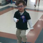 little boy with Down syndrome wearing a blue shirt and brown pants and blue glasses with a pin that says Birthday boy