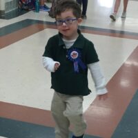 little boy with Down syndrome wearing a blue shirt and brown pants and blue glasses with a pin that says Birthday boy little boy with Down syndrome wearing a blue shirt and brown pants and blue glasses with a pin that says Birthday boy
