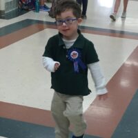 little boy with Down syndrome wearing a blue shirt and brown pants and blue glasses with a pin that says Birthday boy