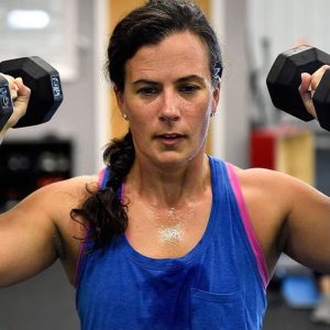 Shawn Cheshire, a white woman with long brunette hair in a ponytail with a concentrating look, at the gym wearing a blue tanktop sweating lifting two fifteen pound dumbells to by her head.