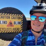 Shawn Cheshire in arizona with her bike helmet blue shirt and red reflective sunglasses with the reflection of her taking a selfie. Shawn is smiling and in the background is the tire from her vehicel with a sign that says Caution Blind Cyclists Ahead. the desert is in the background.
