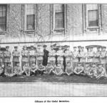 Black and white picture of boys, ages 10 to 18, in military dress in front of a building