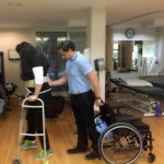 justine using her walker and getting help with her therapist who is standing and has her wheelchair behind him
