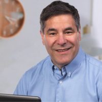 Sheldon Lewis sitting in front of a computer screen wearing a light blue business shirt smiling at the camera