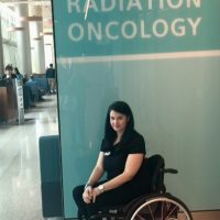 justine sitting in her wheelchair under a sign that says radiation oncology justine sitting in her wheelchair under a sign that says radiation oncology
