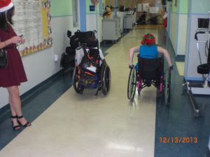 Stephanie and a young man racing wheelchairs down the hallaway
